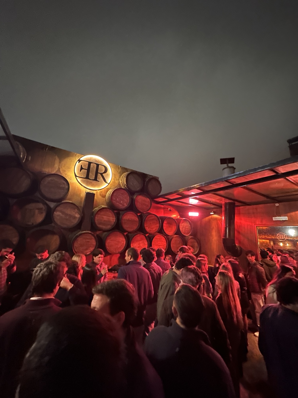Patio de Rancho Bar lleno de gente, con barricas y el emblema iluminado del local